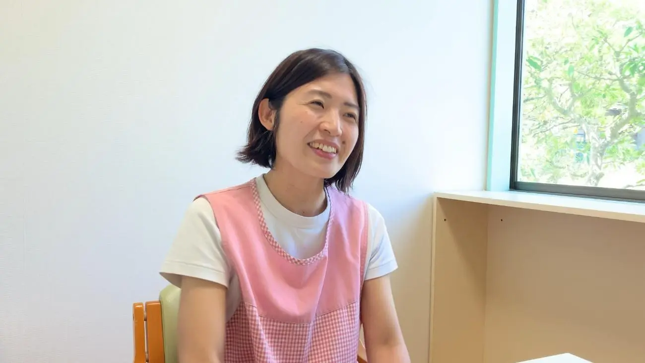 窓際で微笑むピンクのエプロンの保育士