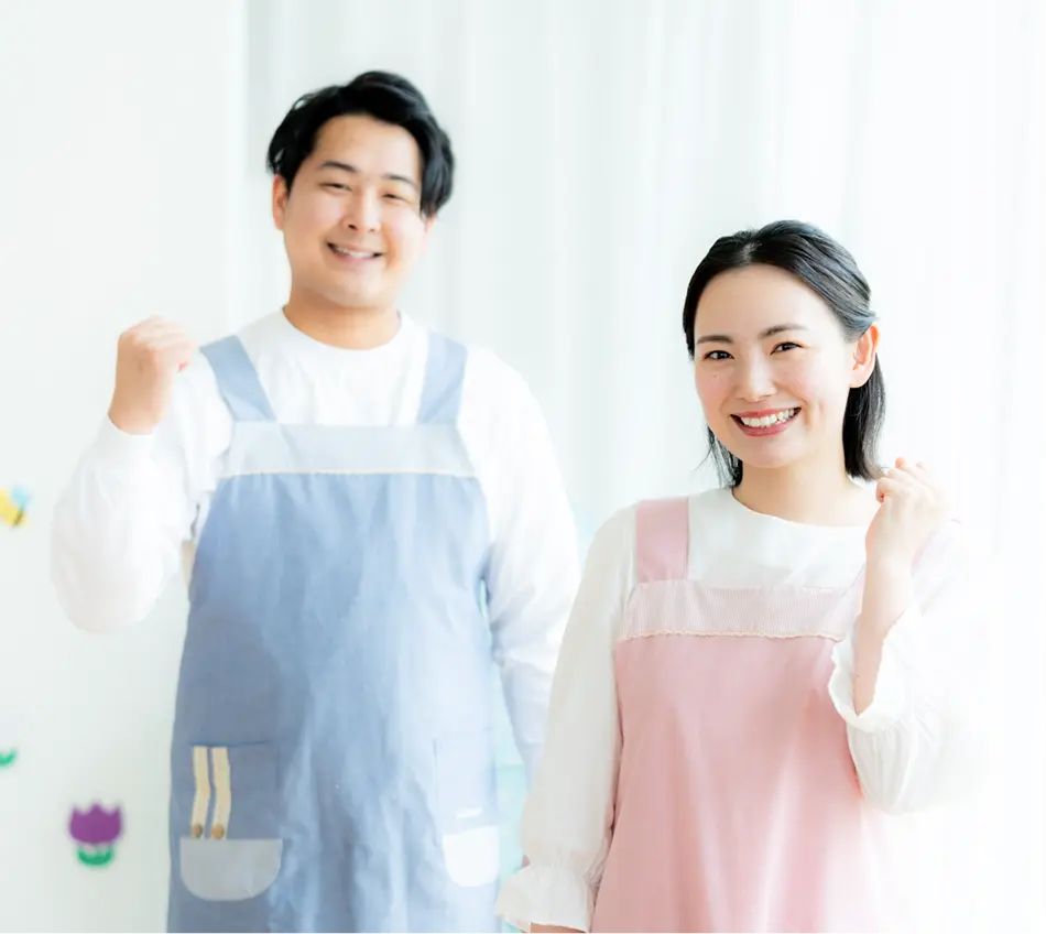エプロン姿で笑顔を見せる男性と女性の保育士、明るい室内背景