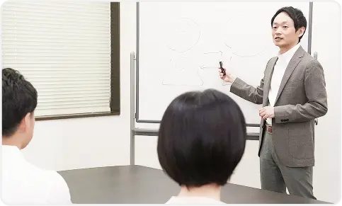 ホワイトボードの前で講義する講師とそれを聞く受講者たち